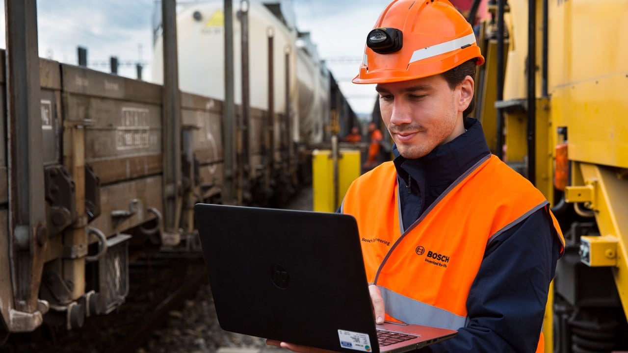 Worker standing between freight trains looking at his laptop.